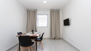 A small, modern room with a dining table, chairs, a window with curtains, and a flat-screen TV mounted on the wall.