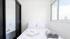 A hotel room with a bed, white linens, a window with a view of buildings, and a mirror reflecting the bed.