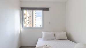 A minimalist hotel room with a bed, two pillows, a window with a view of buildings, and a pair of towels on the floor. - Apto 502 - 2 dorm em Santana (São Paulo)
