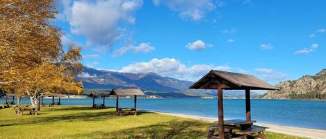 A lakeside park with picnic tables and gazebos, surrounded by mountains.