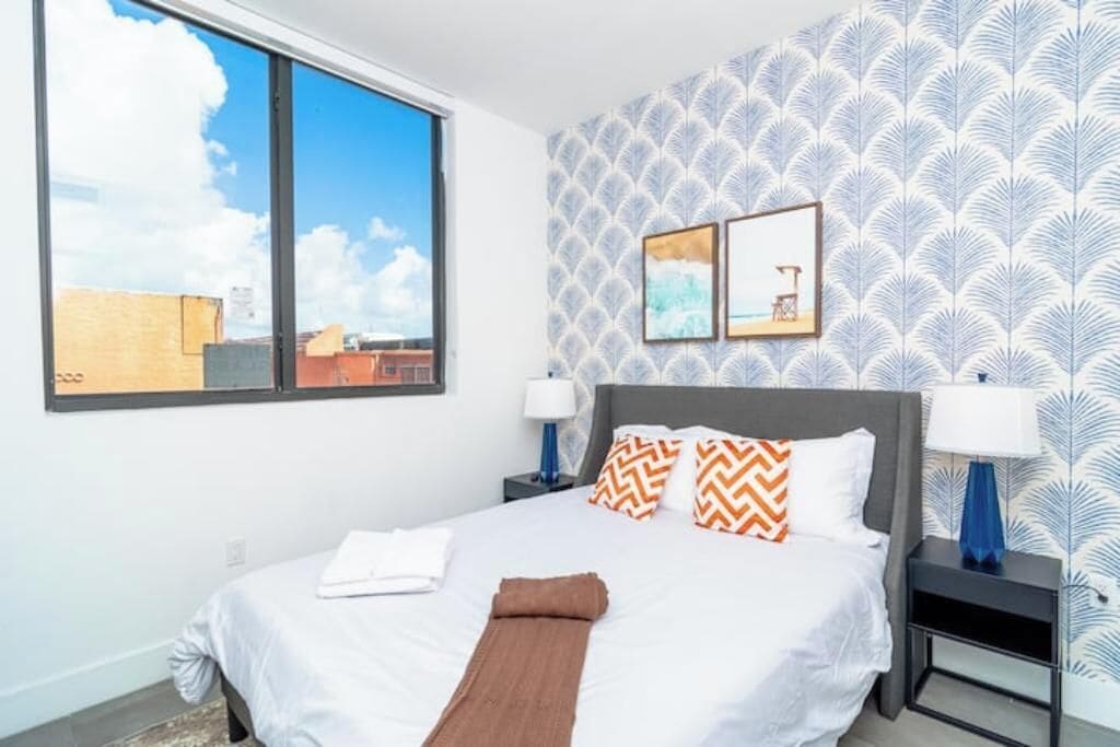 Une chambre d’hôtel avec un grand lit, deux tables de chevet, une fenêtre donnant sur la ville et un papier peint à motifs.