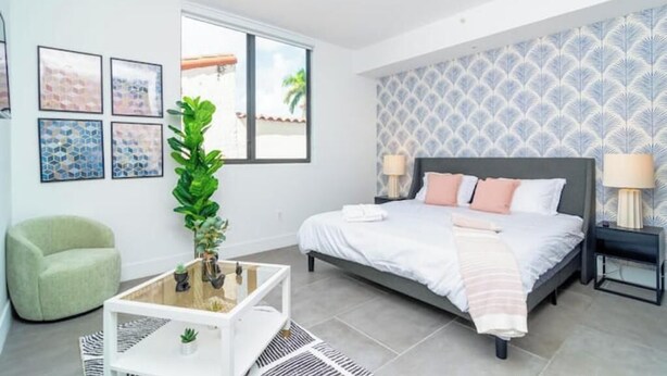 Un dormitorio moderno con una cama, mesitas de noche, un sillón verde, una mesa de centro y un ventanal grande.