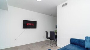 Una sala de estar moderna con un televisor de pantalla plana montado en la pared que muestra 'NETFLIX', un sofá azul y un comedor con sillas.