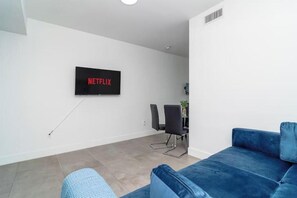 Een moderne woonkamer met een flatscreen-tv aan de muur waarop 'NETFLIX' staat, een blauwe bank en een eethoek met stoelen.