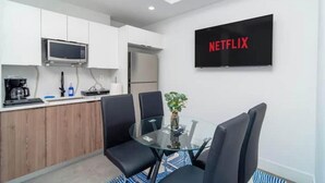 A modern kitchen with white cabinets, a microwave, and a refrigerator. There is a glass table with chairs and a plant. A TV on the wall displays "NETFLIX".