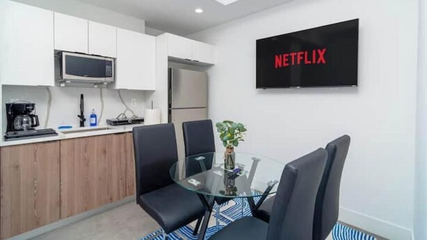 A modern kitchen with white cabinets, a microwave, and a refrigerator. There is a glass table with chairs and a plant. A TV on the wall displays "NETFLIX".