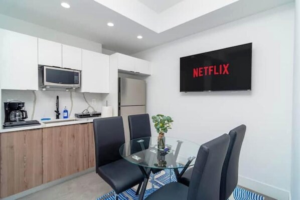Eine moderne Küche mit weißen Schränken, einer Mikrowelle und einem Kühlschrank. Ein Glastisch mit Stühlen und einer Pflanze steht dort. Ein Fernseher an der Wand zeigt "NETFLIX".