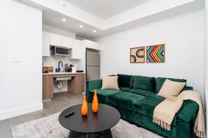Un salon moderne avec un canapé vert, une table basse ronde et un coin cuisine à l’arrière-plan.
