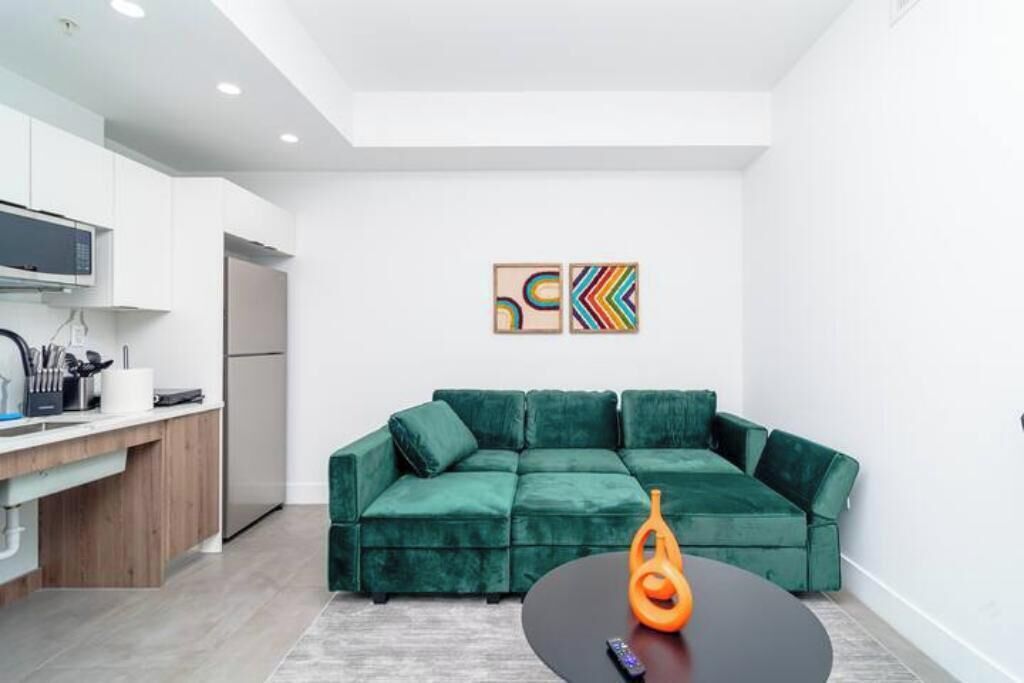 Une cuisine moderne avec des meubles blancs, un four à micro-ondes et un réfrigérateur. On y trouve un canapé vert avec une table basse noire et un objet décoratif orange. Deux tableaux abstraits ornent le mur.