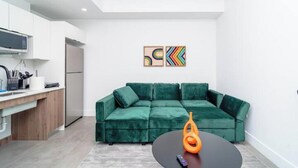 Une cuisine moderne avec des meubles blancs, un four à micro-ondes et un réfrigérateur. On y trouve un canapé vert avec une table basse noire et un objet décoratif orange. Deux tableaux abstraits ornent le mur.