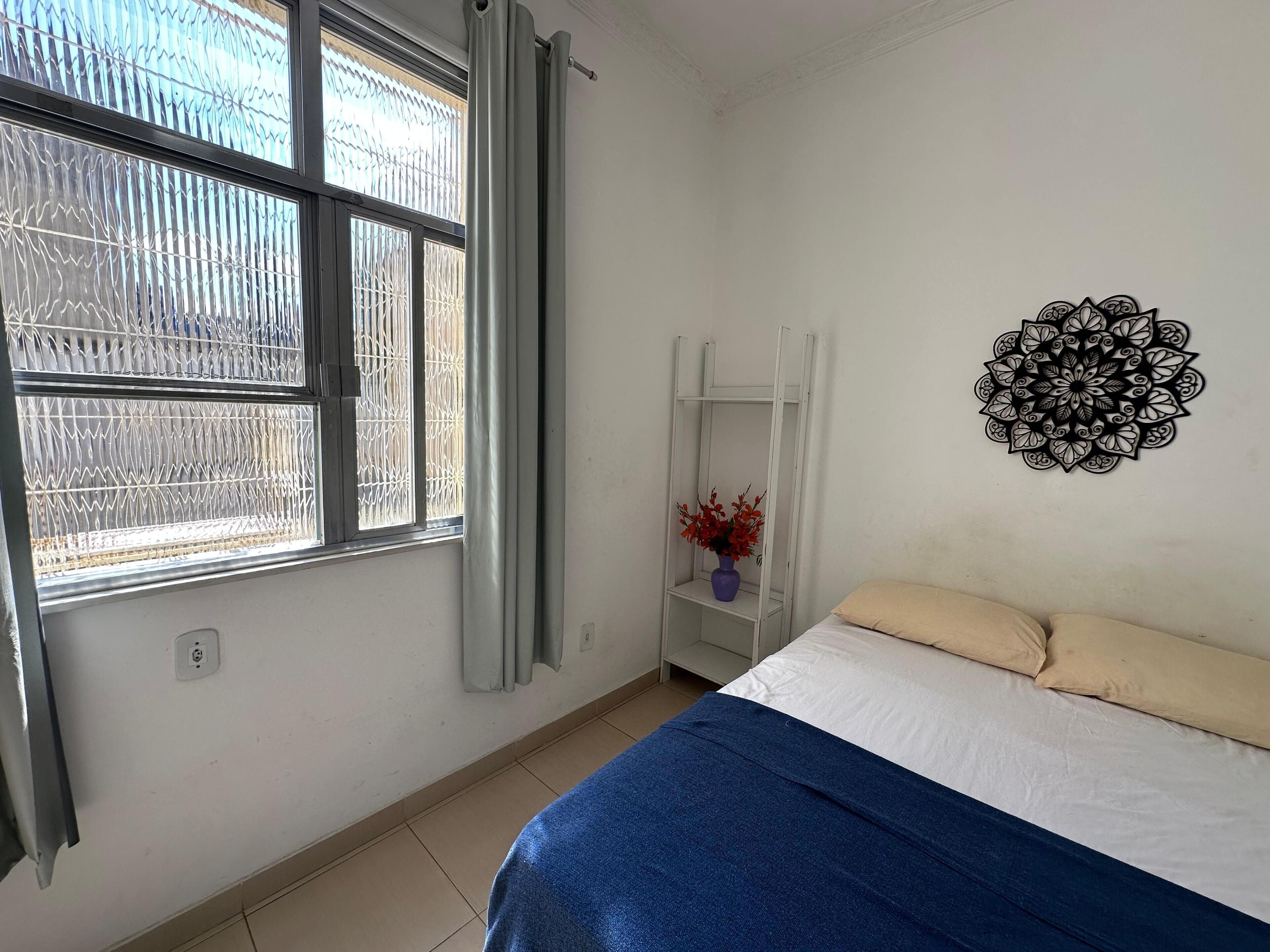A bedroom with a bed, a window with metal bars, a white shelf with a vase, and a wall decoration.