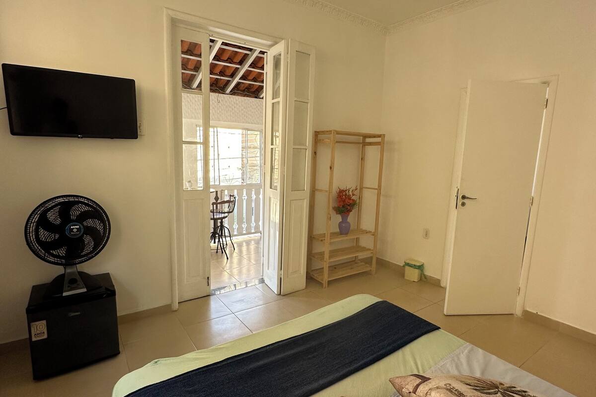 Ein Schlafzimmer mit einem Bett, einem an der Wand montierten Fernseher, einem Standventilator und einem Holzboden mit einer Vase.