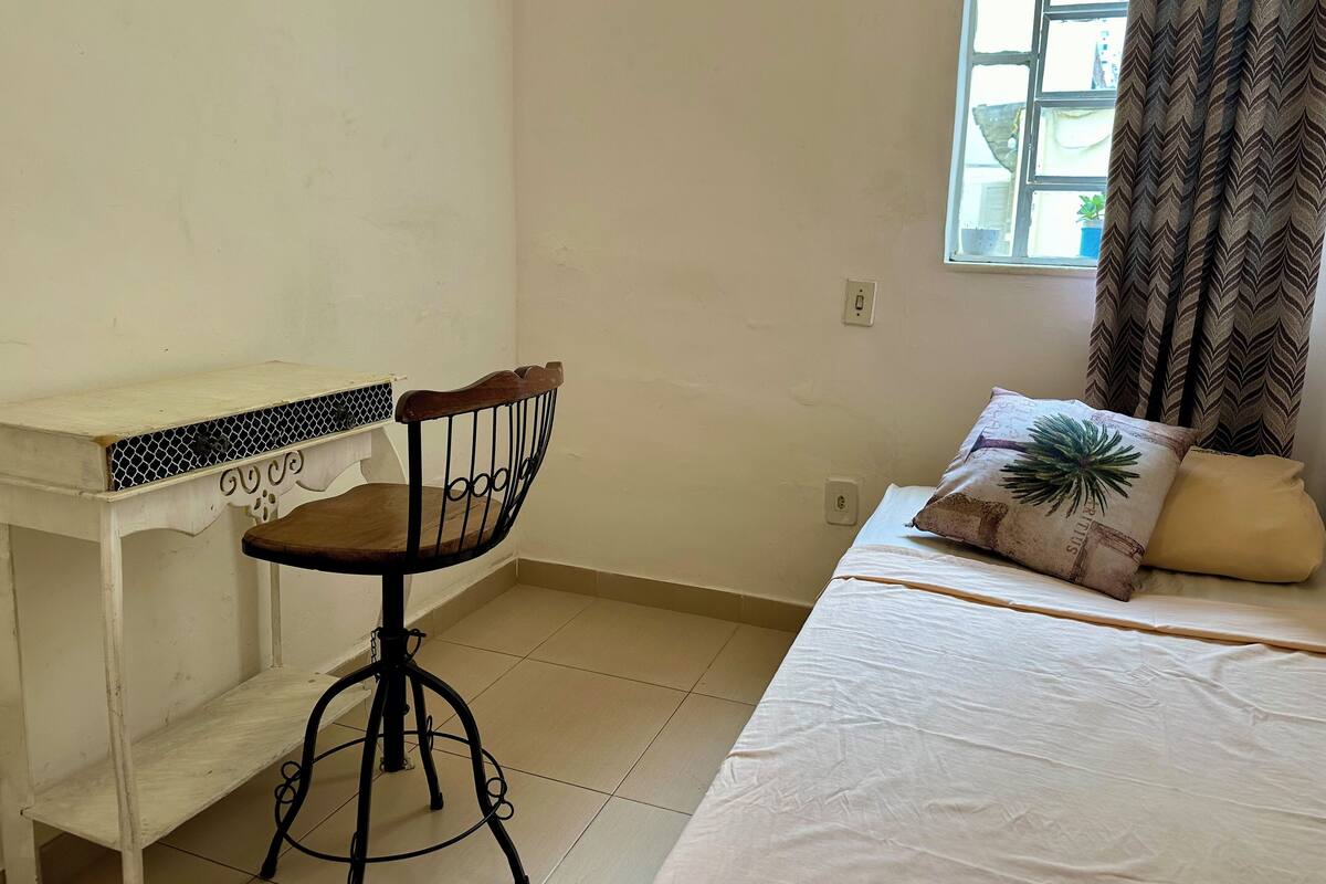 Ein Zimmer mit einem Bett, einem Sessel, einem kleinen Tisch und einem Fenster mit Vorhängen.