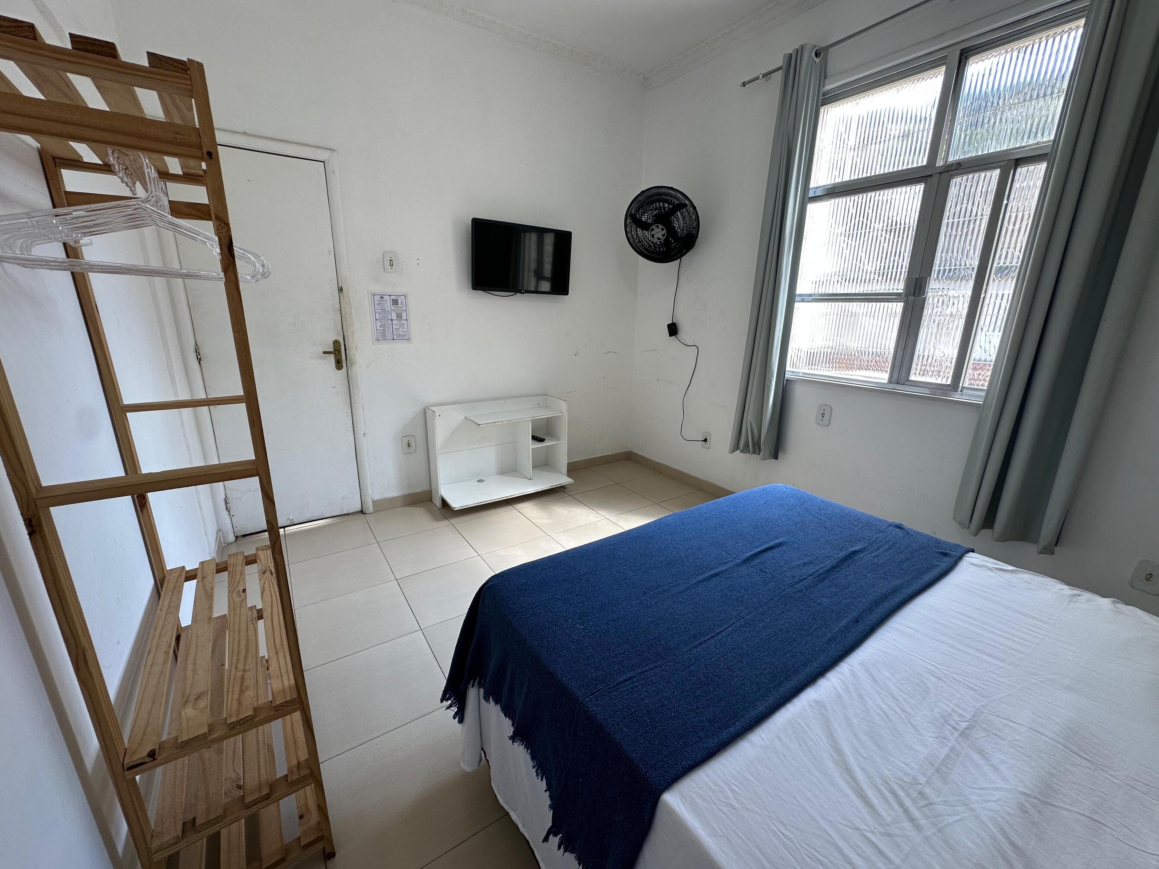A bedroom with a bunk bed, a wooden ladder, a TV on the wall, a window with curtains, and a white shelf unit.