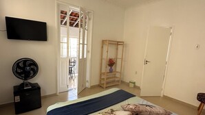 Een slaapkamer met een bed, kussens, een televisie, een ventilator, een deur en een raam.