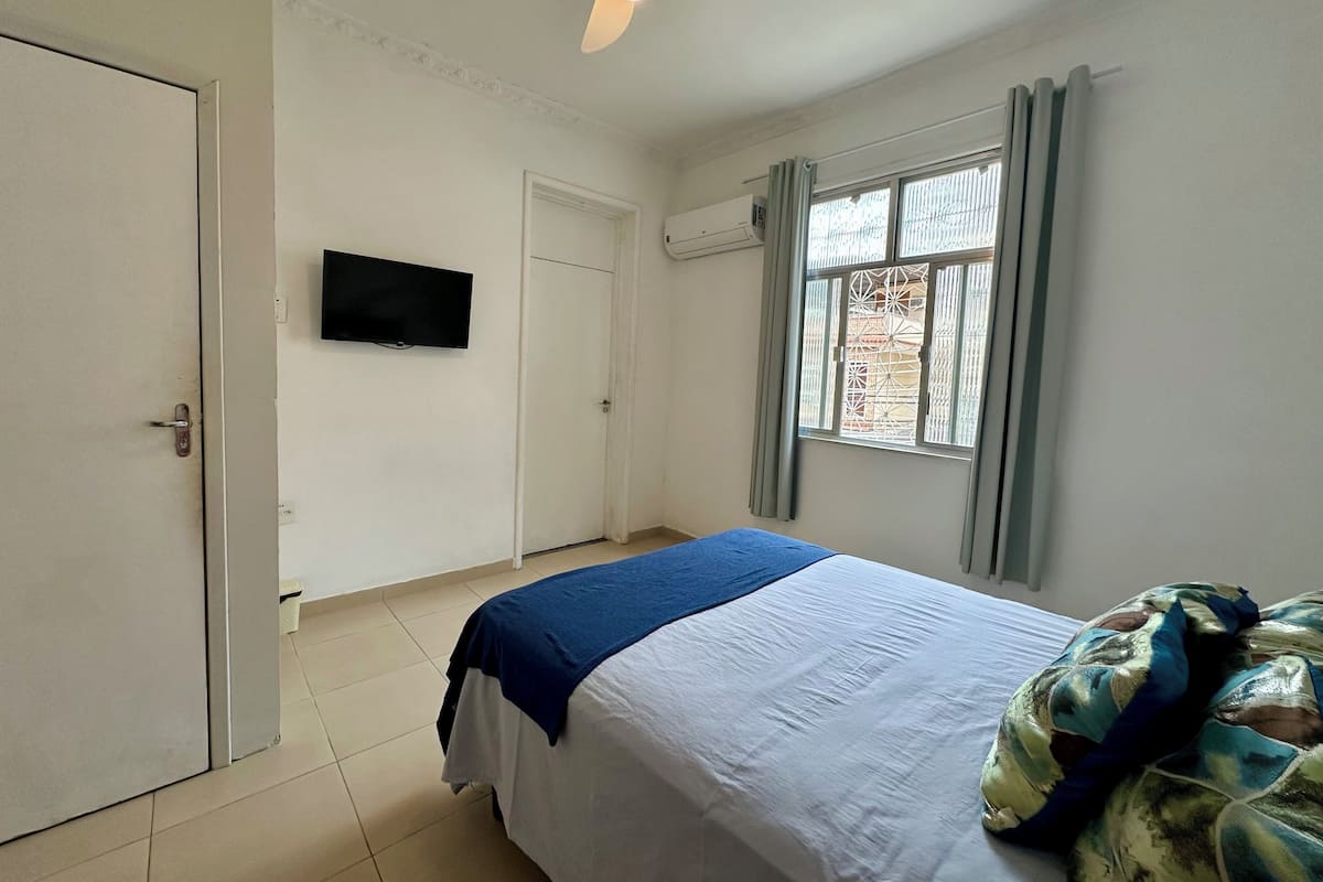 Ein Schlafzimmer mit einem Bett, einem Deckenventilator, einem an der Wand montierten Fernseher und einem Fenster mit Vorhängen.