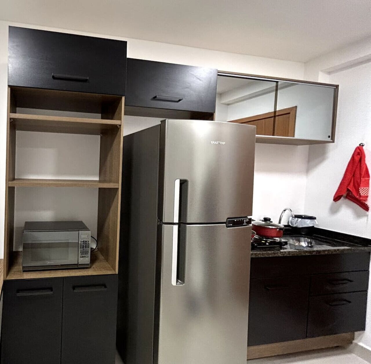 Eine Küche mit einem Edelstahl-Kühlschrank, einer Mikrowelle und einem Herd. Darüber befinden sich Holzregale und ein rotes Handtuch hängt an der Wand.