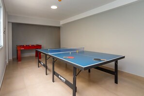 Una mesa de ping pong con dos paletas y una pelota, una mesa de fútbolín roja, y una ventana con persianas.