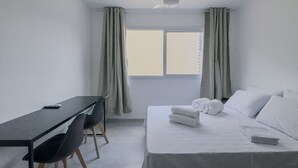 A hotel room with a bed, a desk, and a chair. There is a window with curtains and a ceiling fan. - Studio 521 - sem cozinha (São Paulo)