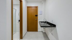 A narrow hallway with a wooden door at the end, a small sink with a mirror, and a light switch on the wall. - Studio 521 - sem cozinha (São Paulo)