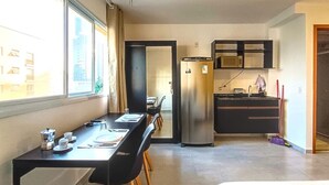 Una habitación de hotel moderna con zona de comedor, kitchenette y un amplio ventanal que proporciona luz natural.