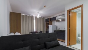 A modern living room with a black sofa, a dining area with a table and chairs, a kitchen with cabinets and a refrigerator, and a wooden door leading to a bathroom.