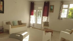 A spacious room with a white sofa, a small table, and a chair. There is a window with curtains and a door with glass panels. - BEAUTIFUL 1ST-FLOOR VILLA APARTMENT (Sainte-Anne)