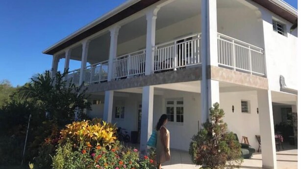 A two-story building with a covered balcony and a garden with colorful flowers. - BEAUTIFUL 1ST-FLOOR VILLA APARTMENT (Sainte-Anne)