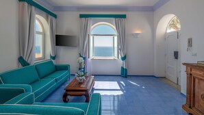 A room with a teal sofa, a wooden coffee table, and a view of the sea through a large window with teal curtains. - AMORE RENTALS - Villa Jacaranda with Pool and Sea View (Praiano)