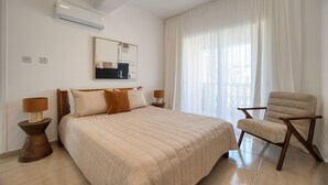 Une chambre avec un grand lit, une chaise, une table de chevet, une lampe, un climatiseur et une fenêtre avec des rideaux légers.
