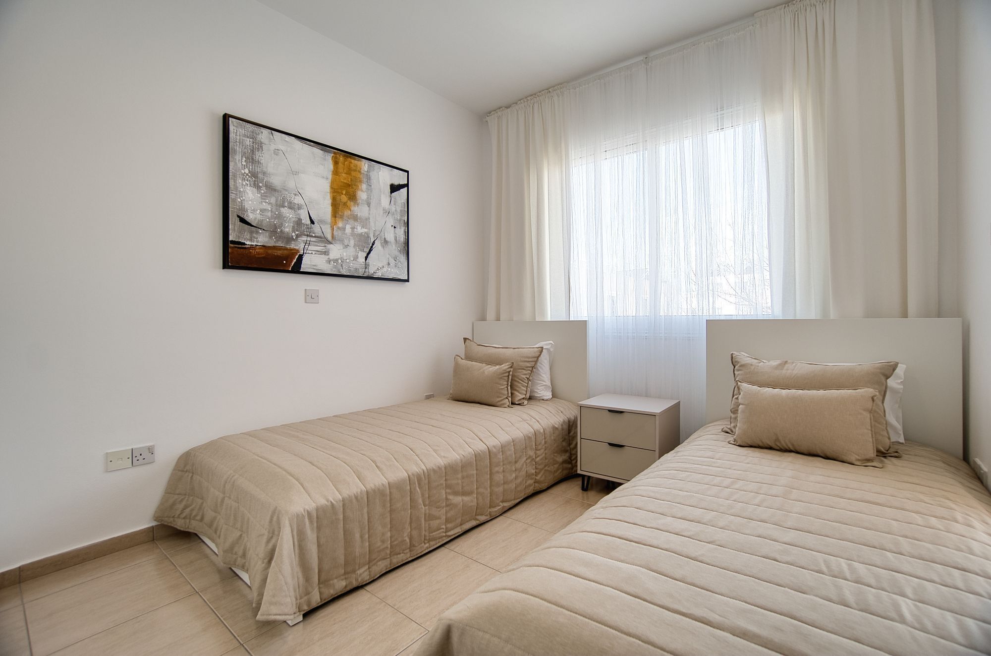Ein Schlafzimmer mit zwei Betten, einem Nachttisch, einem Beistelltisch, einem Gemälde und einem Fenster mit Vorhängen.