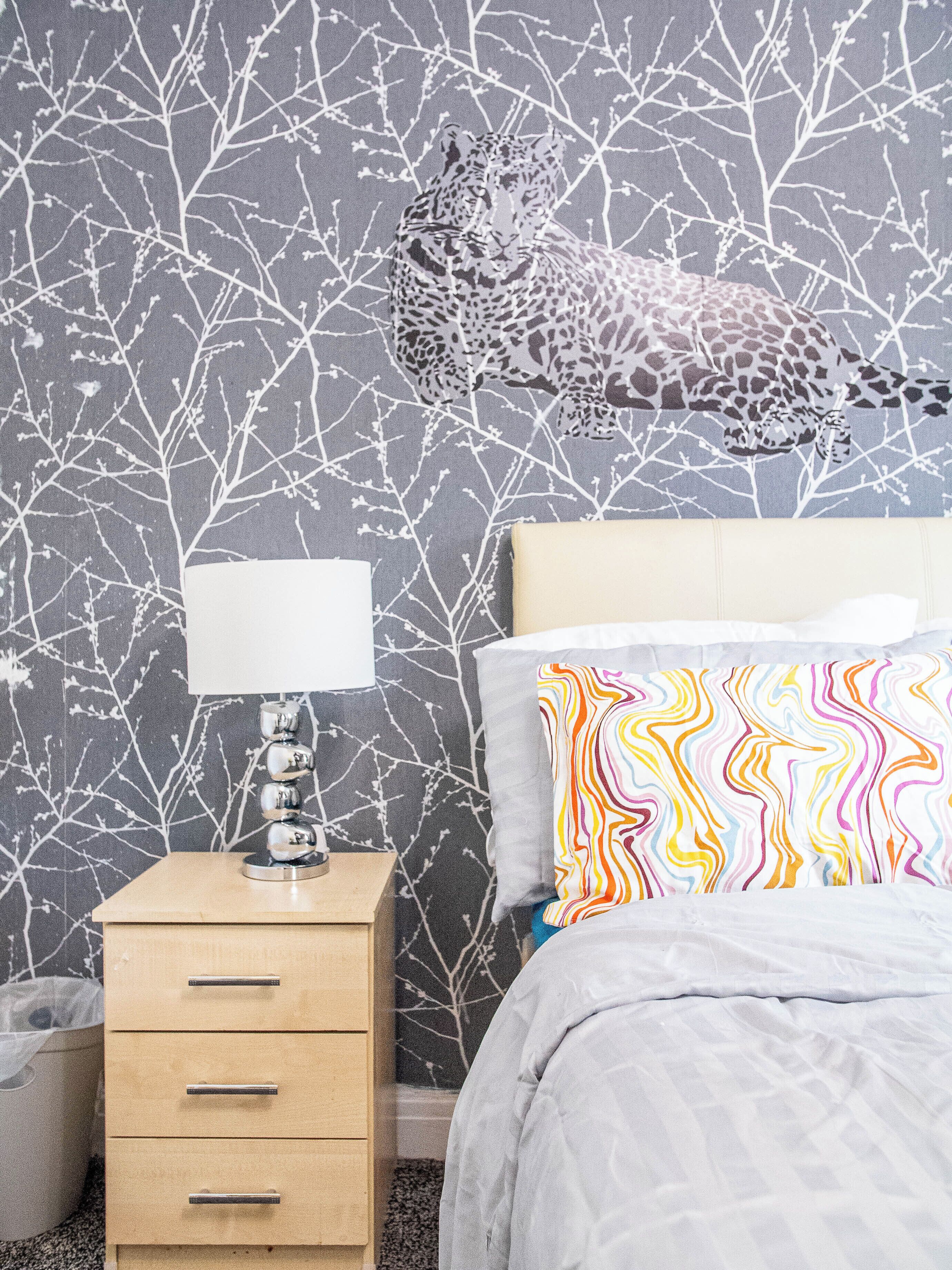 A bedroom with a leopard print wallpaper, a bedside table with a lamp, and a colorful pillow.
