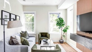A modern living room with a sofa, armchair, coffee table, and a large TV on a wooden cabinet. - Luxe 6-BR + Rooftop Patio | Parking | Metro (Washington)