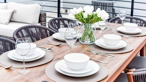 A dining table set with white plates, bowls, and wine glasses, surrounded by wicker chairs.