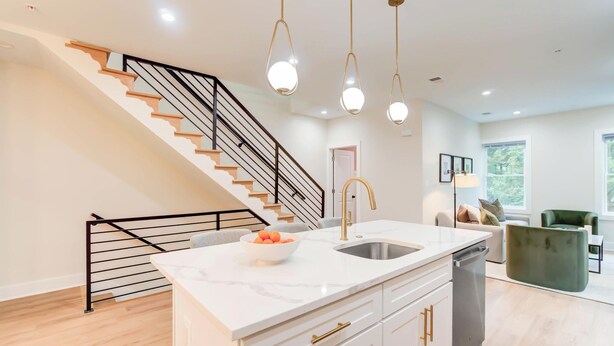 A modern kitchen with a white marble countertop, a stainless steel sink, and a built-in dishwasher. There is a staircase with wooden steps and a black railing leading to another level.