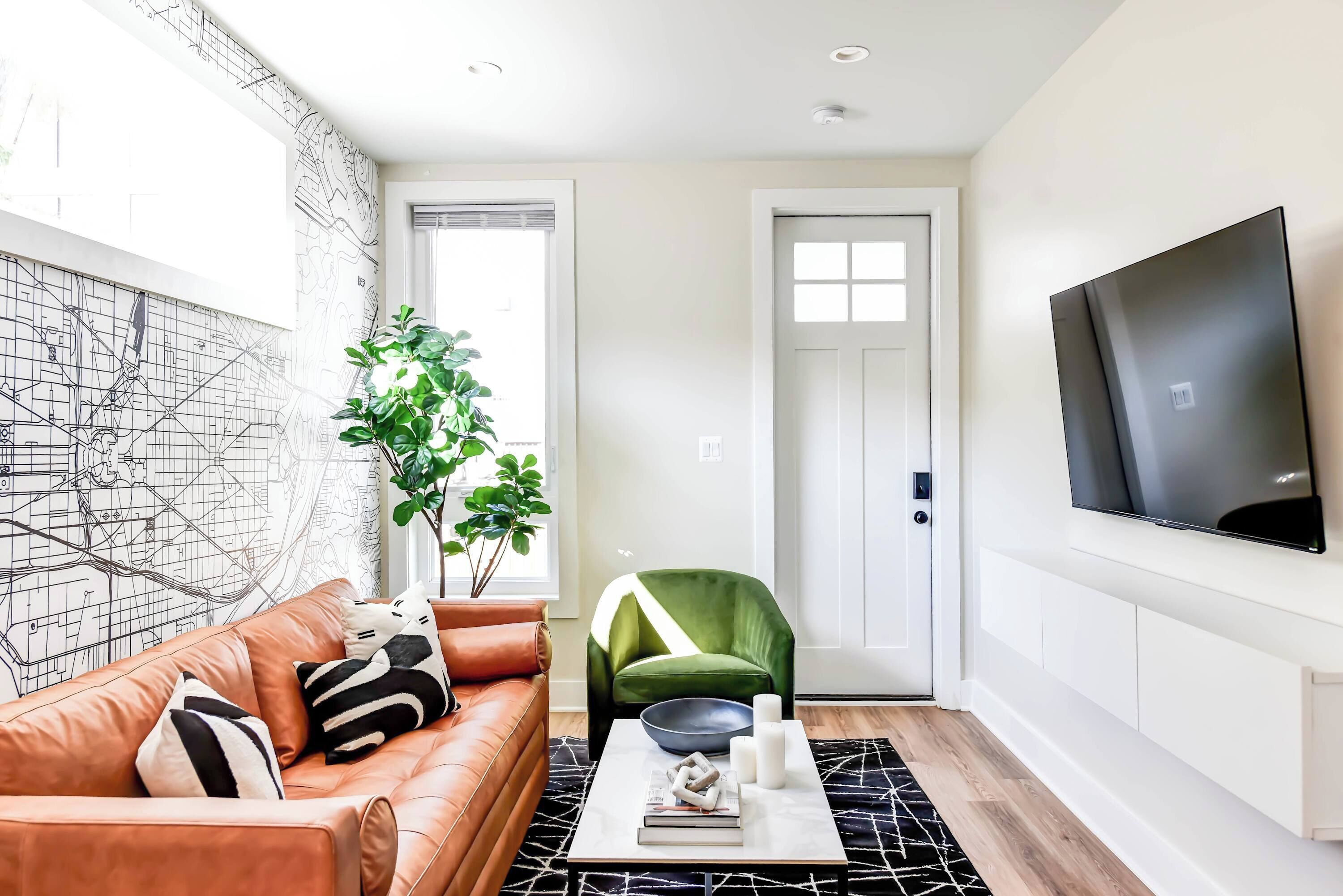 A modern living room with a leather sofa, a green armchair, a coffee table with books and candles, a large TV, and a map wall.