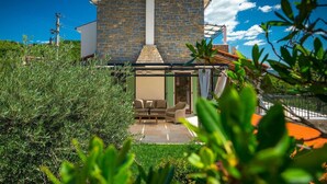 A stone house with a chimney, a covered patio with furniture, and a garden in the foreground.