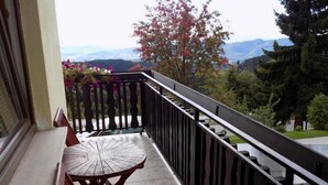En balkon med et rundt bord og stole, med udsigt over et bjergrigt landskab med træer og en klar himmel.