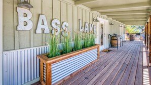 Un porche cubierto con la inscripción "BASS LAKE" en la pared, una maceta de madera con plantas y un barril.