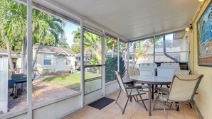 En overdækket terrasse med bord og stole, store vinduer og udsigt til en have med palmer og et hus.