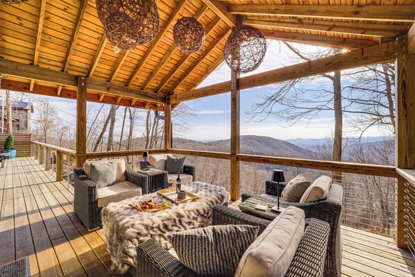 Une terrasse en bois avec des meubles en osier, une table sur laquelle est disposée une assiette de collations, et une vue sur les montagnes.