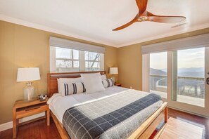 Une chambre à coucher avec un grand lit, une tête de lit en bois, deux tables de chevet avec des lampes, un ventilateur de plafond et de grandes fenêtres offrant une belle vue.