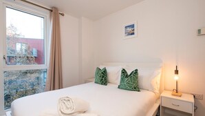 Una cama bien tendida con una toalla enrollada, una mesita de noche con lámpara y una ventana con cortinas.