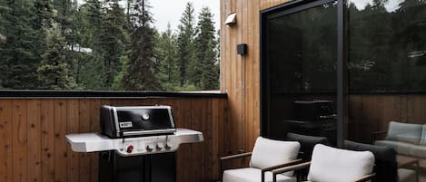 A wooden deck with a Weber grill, outdoor seating, and a large window overlooking a forest.