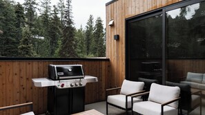 Eine Holzterrasse mit einem Weber-Grill, Sitzgelegenheiten im Freien und einem großen Fenster mit Blick auf einen Wald.