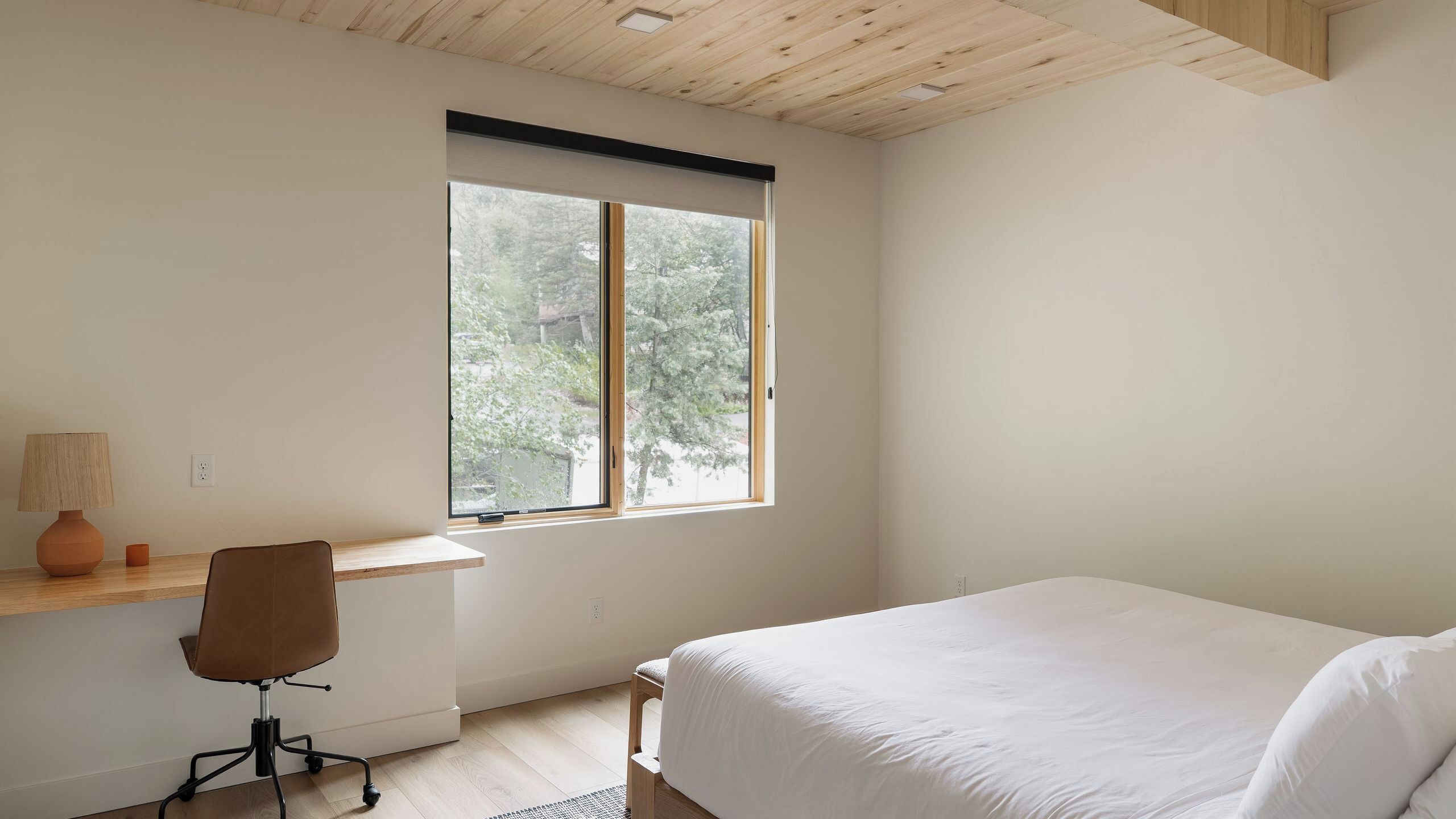 A bedroom with a bed, a desk, a chair, a window, and a ceiling with wooden beams.