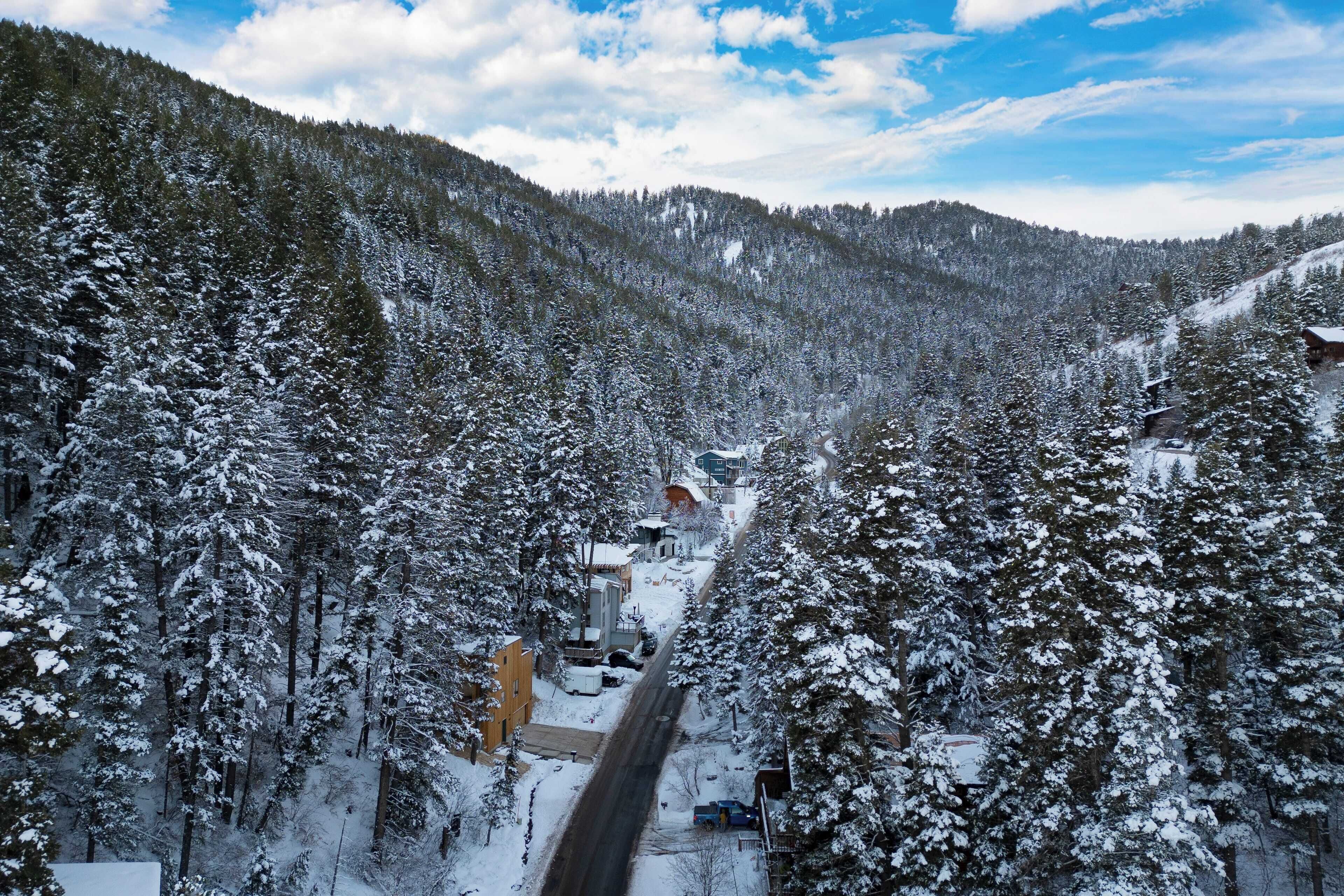Une route de montagne enneigée, bordée d’épais conifères, qui mène à un petit village avec des bâtiments et une route bien visible.