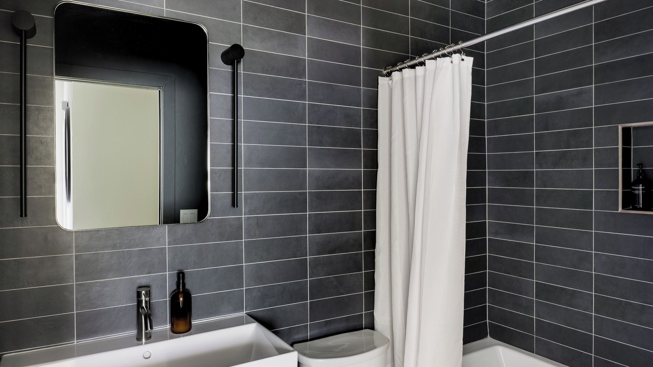 A modern bathroom with dark tiled walls, a white bathtub, a toilet, a mirror, and a shower curtain.