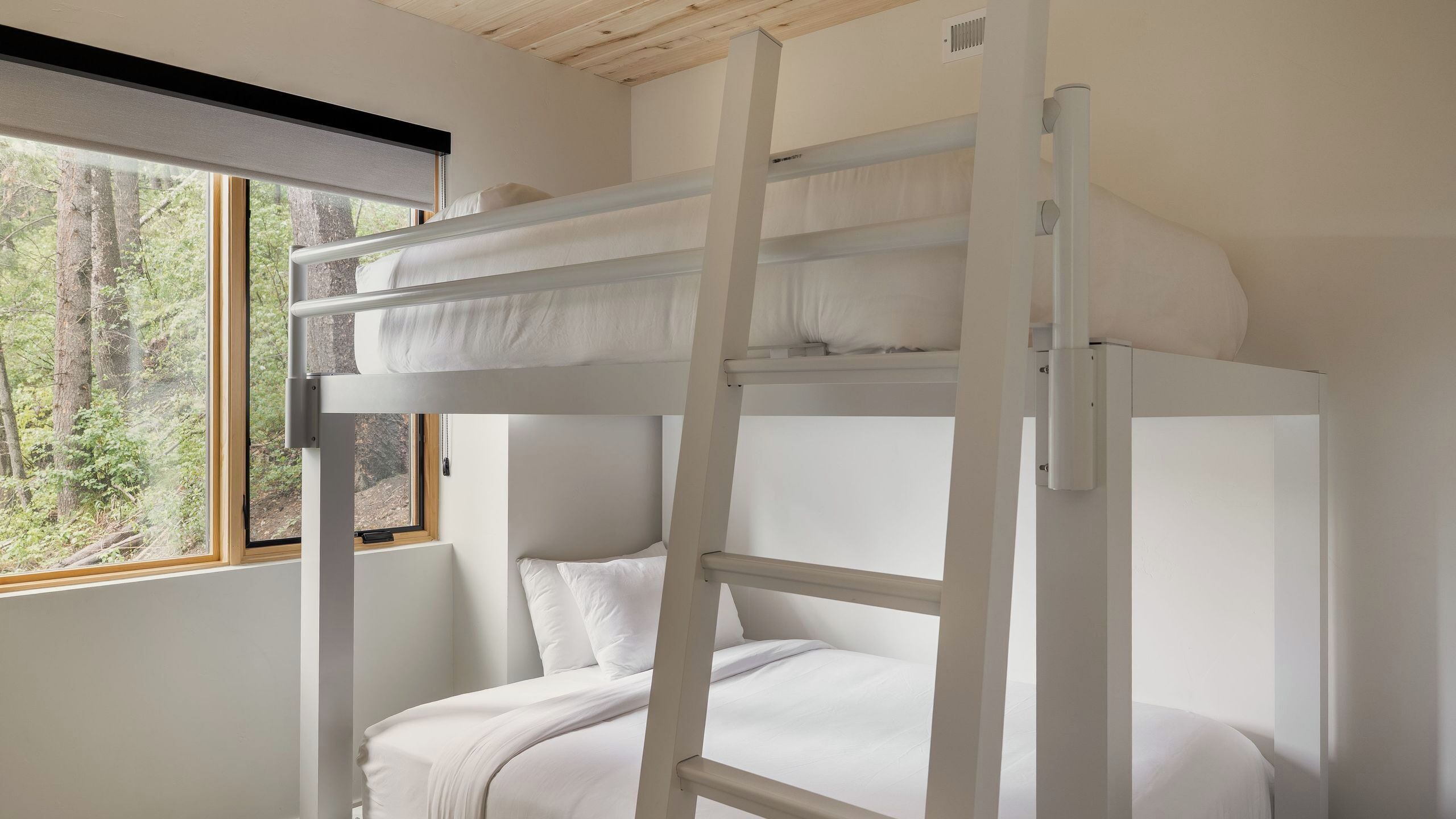 Una cama litera con ropa de cama blanca, una ventana con vistas a árboles y un techo de madera.
