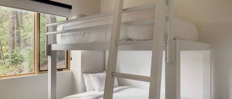 Una cama litera con ropa de cama blanca, una ventana con vistas a árboles y un techo de madera.